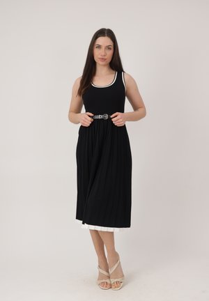 Kobieta poprawia czarną sukienkę midi z paskiem, białą lamówką i warstwowym dołem, ma na sobie beżowe sandały na obcasie, stoi na tle jasnego, jednolitego tła.