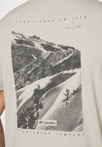 Světle šedé tričko vyrobené z bavlny, s černobílou grafikou horské krajiny a logem "Columbia Sportswear Company" ve spodní části.