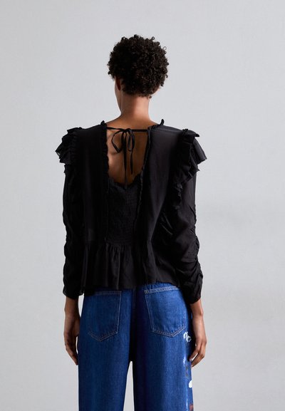 Black blouse with ruffled sleeves, open back tied with ribbon, textured design. Paired with blue denim pants featuring colorful embroidery.