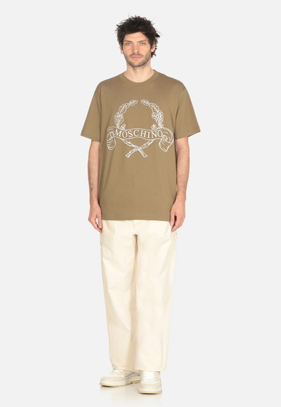 T-shirt verde oliva con stampa e design grafico bianco; abbinata a pantaloni beige a grandezza libera e sneakers di colore chiaro.