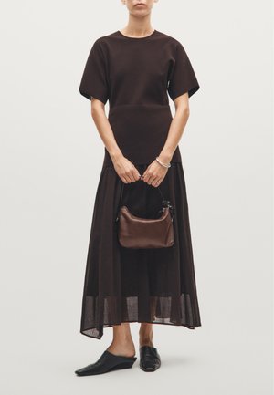 Maglietta corta marrone a maniche corte e una lunga gonna marrone a strati. Tiene una piccola borsa di pelle marrone, indossa mule neri. Tessuti morbidi e design fluido.