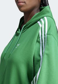 Persoon met een groene hoodie met witte strepen op de schouders en trekkoorden, zichtbaar van de nek tot halverwege de romp.