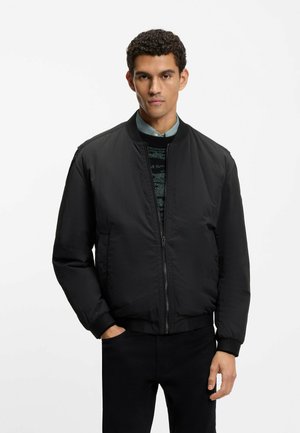 Hombre con chaqueta bomber negra sobre un suéter estampado y camisa con cuello, de pie con una mano en el bolsillo contra un fondo liso.