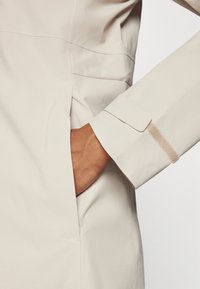 Beige lätt jacka med en synlig sidoficka och en ärm med kardborrestängning, tillverkad av mjukt, strukturerat tyg.