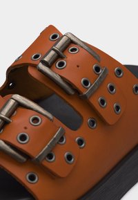 Κοντινή λήψη ενός πορτοκαλί δερμάτινου σανδαλιού με δύο ρυθμιζόμενες μεταλλικές αγκράφες και πολλαπλές μεταλλικές τρύπες σε μαύρη σόλα.