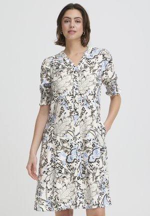 Ženska v kolenkah dolgi cvetlični obleki z gumbi, z napihnjenimi kratkimi rokavi in žepi, stoji pred enostavnim ozadjem.