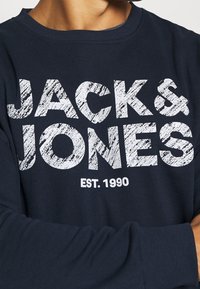 Marinblå sweatshirt med rundad halsringning, prydd med stora vita texter "JACK & JONES" och "EST. 1990" i en texturerad, sliten stil.
