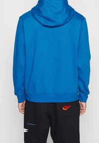 Person i blå hoodie och svarta Nike sweatpants med röd logga, visad från baksidan mot en enkel bakgrund.