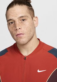 Jachetă cu fermoar Nike în roșu, cu accente bleumarin, guler ribbed și un logo alb. Material neted, cu un design ajustat și închidere cu fermoar.