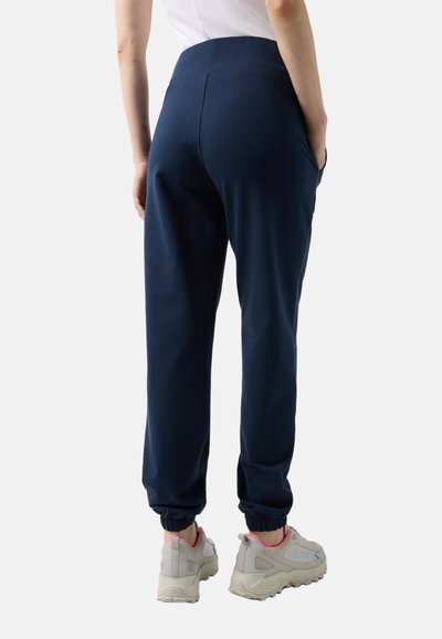 Person trägt marineblaue Jogginghose und weiße Sneaker, von hinten mit den Händen in den Taschen gezeigt, steht vor einem einfarbigen Hintergrund.