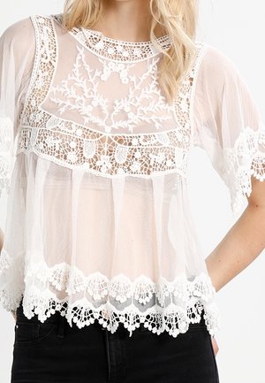 Nainen, joka käyttää valkoista läpikuultavaa pitsipaitaa, jossa on kukkaompeleita, aaltoilevat reunat ja väljät lyhyet hihat, yhdistettynä mustiin housuihin.