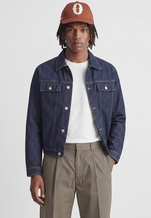 Jeune homme avec des dreadlocks portant une casquette marron, une veste en jean bleu foncé, un t-shirt blanc et un pantalon beige, debout devant un fond uni.