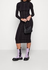 Schwarzes, figurnahes Kleid mit grünen, vertikalen Nähten und gewellten Bündchen, kombiniert mit einer kleinen schwarzen Handtasche und lavendelfarbenen, schwarzen klobigen Stiefeln.