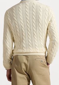 Kremowy sweter w warkoczowy splot z prążkowanymi mankietami i ściągaczem, zestawiony z jasnobeżowymi spodniami ozdobionymi małą kieszonką i guzikiem.