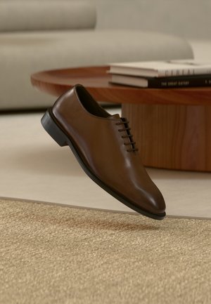 Zapato oxford de cuero marrón con suela negra suspendido sobre una alfombra beige tejida, con una mesa de madera y libros apilados al fondo.