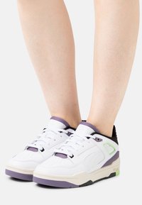 White sneakers with purple accents, featuring a textured leather upper, flat laces, perforated toe, and a contrasting rubber sole.