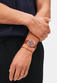 Brown leather strap watch with a dark brown round face. The watch features gold-tone accents and a minimalist design, worn on a hand.