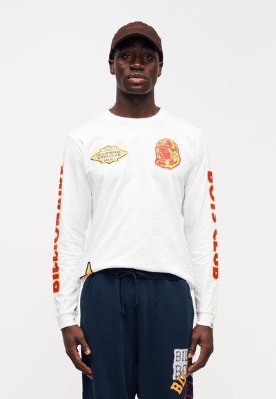 Άσπρο πουκάμισο με μακριά μανίκια, διακοσμημένο με κόκκινα γραφικά, συμπεριλαμβανομένου ενός αστροναύτη και κειμένου. Συνδυάζεται με ναυτικό μπλε φόρμας παντελόνια με λογότυπα.