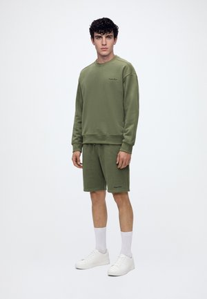 Giovane uomo che indossa una felpa verde oliva e pantaloncini abbinati con calzini bianchi e scarpe da ginnastica bianche, in piedi su uno sfondo semplice.