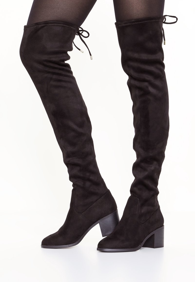 Bottes noires en daim hautes avec des talons carrés, portées par-dessus des collants noirs, arborant des cordons de serrage à l'arrière supérieur de chaque botte.