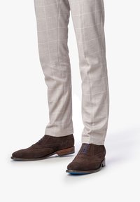 Scarpe da uomo in suede marrone con dettagli brogue e lacci blu, abbinate a pantaloni a quadri leggeri grigi. Le scarpe presentano un tacco in legno.
