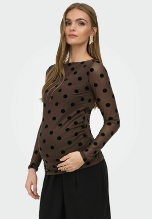 Gravid kvinne med langt hår iført gjennomsiktig brun langermet topp med svarte polkadotter og svarte bukser, som forsiktig holder magen sin.