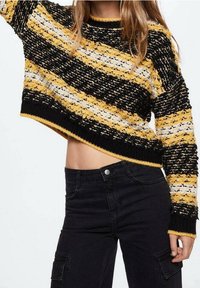 Maglione corto all'uncinetto con strisce nere, gialle e bianche. Presenta un colletto rotondo e un bordo a costine. Indossato con pantaloni neri a vita alta.