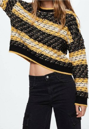Suéter de crochet cortado con rayas negras, amarillas y blancas. Presenta un escote redondo y un dobladillo acanalado. Combinado con pantalones negros de tiro alto.