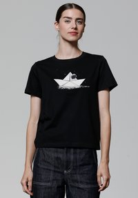 Zwart katoenen T-shirt met een witte afbeelding van een papieren boot met een astronaut binnenin, tegen een sterrenachtige achtergrond. Korte mouwen, ronde halslijn.