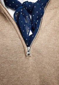 Beige stickad tröja med en fjärdedels dragkedja och en texturerad yta, lagd över en vit skjorta, accentuerad med en marinblå scarf med prickar.