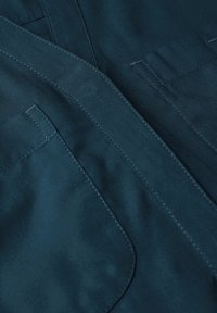 Tessuto teal scuro con una texture liscia. Presenta cuciture visibili e due tasche ampie. Il design include un colletto pieghettato e una apertura frontale.