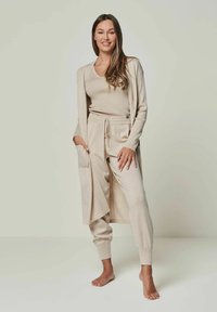 Beiges Loungewear-Set, bestehend aus einer langen Strickjacke, einem eng anliegenden Oberteil und Jogginghosen mit elastischen Bündchen und einem Kordelzugbund, aus weichem Material gefertigt.