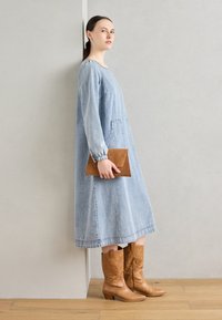Jeanskleid mit langen Ärmeln, seitlichen Taschen, blauer Farbe und einem knielangen Saum. Kombiniert mit braunen Lederstiefeln bis zum Knie und einer braunen Clutch.