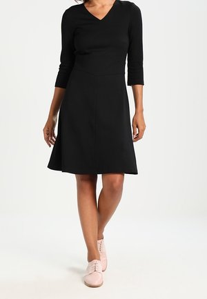 Woman walking wearing a knee-length black dress with three-quarter sleeves and light pink lace-up shoes against a plain background.