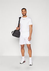 Hvit atletisk t-skjorte med svart logo, matchende hvite shorts og atletiske sko med svarte detaljer. Svart treningsbag med skulderrem.