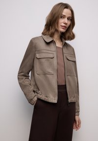 Brun, tekstureret jakke med lynlås foran, krave og to brystlommer. Har elastiske ærmekanter og linning for et tætsiddende look.