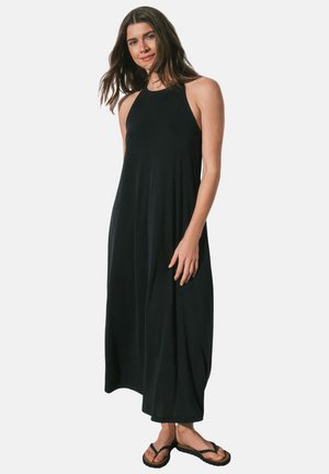 Donna con capelli castani lunghi che indossa un abito maxi nero senza maniche e infradito nere, in piedi davanti a uno sfondo chiaro e semplice.