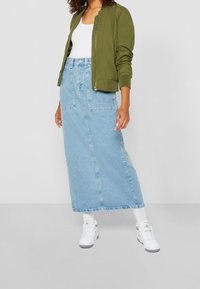 Veste bomber vert olive, débardeur blanc, jupe maxi en denim bleu clair, baskets blanches et chaussettes blanches. La jupe a des poches avant et des coutures.