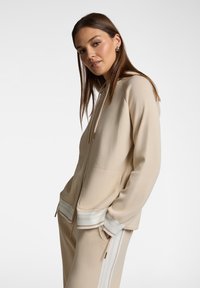 Felpa con zip beige realizzata in morbido tessuto, con cappuccio foderato, tasche frontali e dettagli a costine a righe sull'orlo. Texture liscia e design aderente.