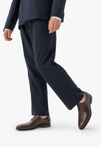 Hnědé kožené společenské boty s kulatou špičkou a šněrováním, doplněné tmavě modrými upravenými kalhotami, které mají hladkou texturu.