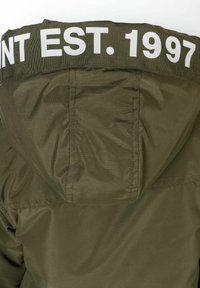 Olivgrön jacka med texturerad yta, med huva och ett vitt band med texten "EST. 1997" över ryggen.