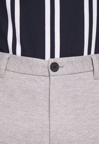 Lichtgrijze broek van gestructureerde stof, met een zwarte knop, met een marineblauw gestreept shirt dat gedeeltelijk zichtbaar is bovenaan.