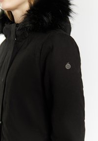 Sort vinterjakke med en fyldig sort faux fur krave, lynlåslukning og et lille emblem på ærmet, der viser detaljeret håndværk.