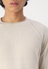 Pull en tricot beige avec un motif texturé et des rayures horizontales. L'encolure arrondie et les manches raglan mettent en valeur le design.