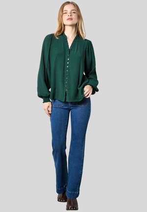 Eine Frau steht in einer dunkelgrünen, knopfgebenden Bluse mit Puffärmeln, blauen Jeans und braunen, mit Nieten besetzten Stiefeletten vor einem grauen Hintergrund.