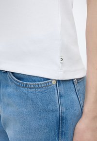 Κοντινό πλάνο ενός ατόμου που φοράει λευκό ριμπ πουκάμισο και μπλε τζιν, με έμφαση στη λεπτομέρεια του τελειώματος του πουκαμίσου και της τσέπης του τζιν.