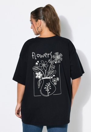 Vrouw met een paardenstaart die een zwart oversized T-shirt draagt met een witte lijntekening van bloemen in een vaas en het woord "flowers" op de rug.