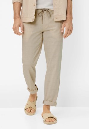 Mann trägt hellbeige Kordelzughose, an den Bündchen hochgekrempelt, kombiniert mit beigen Sandalen mit überkreuzten Riemen, stehend vor einfachem Hintergrund.