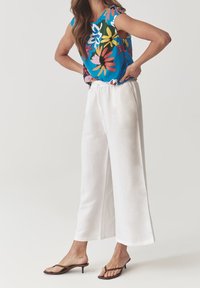 Blusa a maniche corte floreale blu con motivi multicolori, abbinata a pantaloni bianchi larghi in lino e sandali con tacco e cinturini marroni.