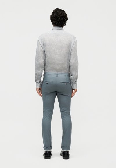 Άνδρας με σγουρά μαλλιά, ντυμένος με ανοιχτό γκρι πουκάμισο μακρυμάνικο που έχει φορεθεί μέσα σε ανοιχτό μπλε στενά παντελόνια και μαύρα παπούτσια, στεκεται με την πλάτη προς τα εμάς.